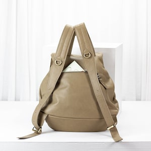 May include: A tan leather backpack with a zippered pocket on the front. The backpack has two adjustable straps and a top handle.