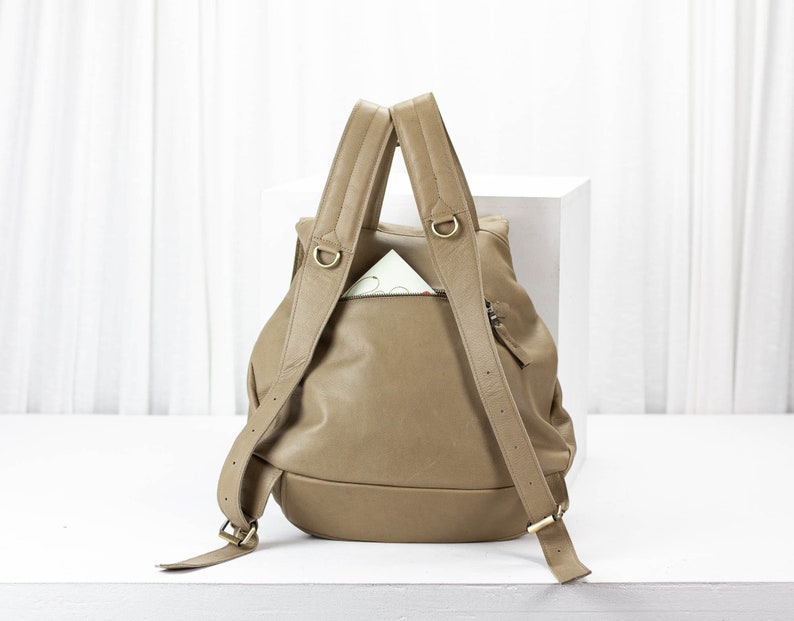 May include: A tan leather backpack with a zippered pocket on the front. The backpack has two adjustable straps and a top handle.