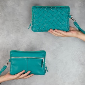 Puede incluir: Dos bolsos de muñeca de cuero turquesa. Un bolso presenta un diseño tejido, mientras que el otro tiene una superficie lisa con un bolsillo frontal con cremallera. Ambos bolsos tienen una correa de muñeca y herrajes plateados. El fondo es gris sólido.