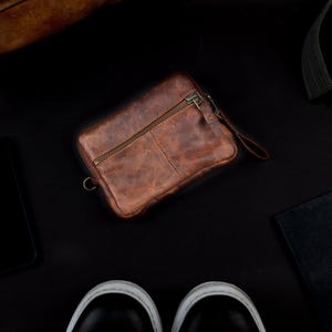 May include: A brown leather wallet with a zipper closure and a wrist strap. The wallet is rectangular and has a distressed appearance. A black smartphone, a notebook, a pen, a belt, and a pair of black shoes are also visible on a black surface.