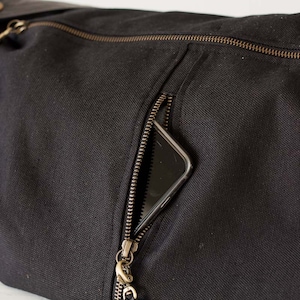 Op de afbeelding: Zwarte canvas tas met een ritsvak, met een goudkleurige rits en hardware. Een smartphone is gedeeltelijk zichtbaar in de zak. De tas heeft een leren riem en metalen knoopdetails.