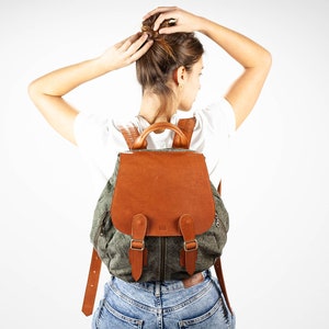 May include: A green canvas backpack with brown leather accents and a flap closure. The backpack has a zipped pocket on the front and adjustable straps.