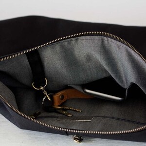 Op de afbeelding: Zwarte canvas messenger bag met een grijze binnenkant. De tas is open en onthult een smartphone, sleutels en een leren sleutelhanger. De rits is messingkleurig. De tas is ontworpen voor het dragen van essentials.