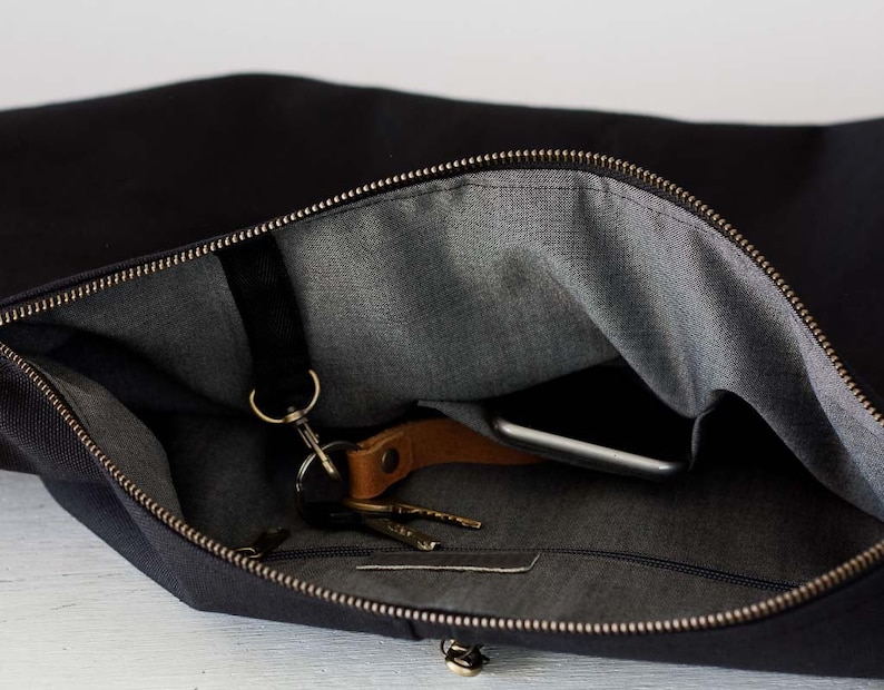 Op de afbeelding: Zwarte canvas messenger bag met een grijze binnenkant. De tas is open en onthult een smartphone, sleutels en een leren sleutelhanger. De rits is messingkleurig. De tas is ontworpen voor het dragen van essentials.