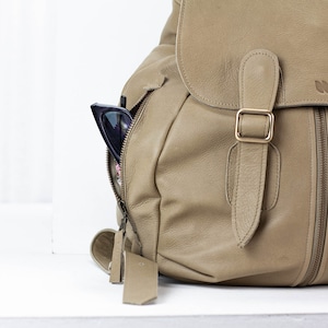 May include: A tan leather backpack with a zippered front pocket and a pair of sunglasses sticking out of the pocket. The backpack has a buckle closure and a leather strap.