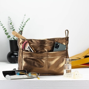 Könnte beinhalten: Ein goldfarbener Lederorganizer mit mehreren Taschen und Fächern. Der Organizer ist gefüllt mit Gegenständen wie einem Make-up-Pinsel, einem Smartphone und einem kleinen Parfümflakon.
