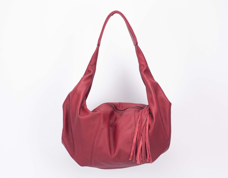 K&ouml;nnte beinhalten: Burgunderfarbene Hobo-Tasche aus Leder mit langem Riemen und Quasten. Die Tasche hat einen Rei&szlig;verschluss und ein weiches, l&auml;ssiges Design. Die Tasche ist aus glattem, geschmeidigem Leder gefertigt.