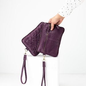 May include: Two purple leather wristlet bags. One has a woven design, and the other has a zipper pocket. Both have wrist straps and gold-colored hardware. The bags are a deep shade of purple.