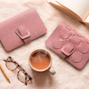 Könnte beinhalten: Zwei rosafarbene Ledergeldbörsen mit Schnallenverschlüssen, eine davon mit geometrischem Design, liegen auf einer flauschigen Oberfläche. Eine Tasse Tee, ein goldener Stift und eine Schildpattbrille vervollständigen die Szene.