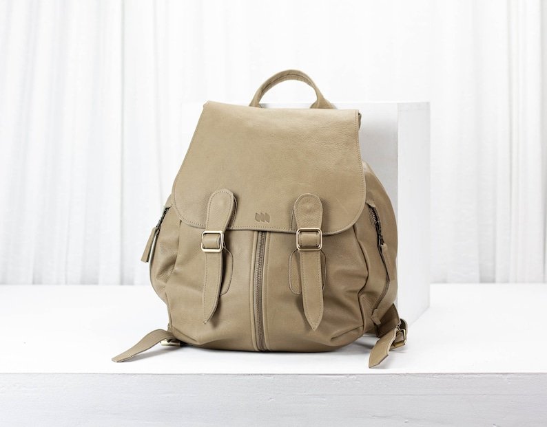 May include: A tan leather backpack with two adjustable straps and a zipper closure. The backpack has a flap with two buckles.