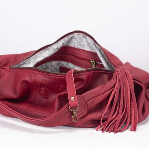 K&ouml;nnte beinhalten: Eine rote Leder-Umh&auml;ngetasche mit langem Riemen und gro&szlig;em Quastenanh&auml;nger. Die Tasche hat einen Rei&szlig;verschluss und ein gemustertes Innenfutter. Die Beschl&auml;ge sind goldfarben.