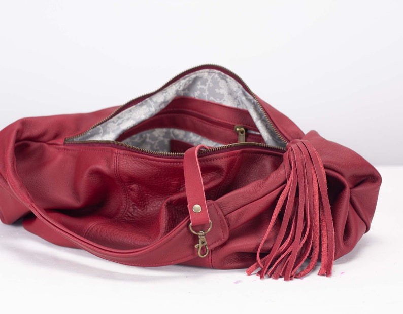 K&ouml;nnte beinhalten: Eine rote Leder-Umh&auml;ngetasche mit langem Riemen und gro&szlig;em Quastenanh&auml;nger. Die Tasche hat einen Rei&szlig;verschluss und ein gemustertes Innenfutter. Die Beschl&auml;ge sind goldfarben.