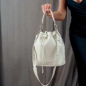 Puede incluir: Un bolso de cubo de cuero blanco con cierre de cordón y una correa larga. El bolso tiene herrajes plateados y un detalle de cadena plateada.