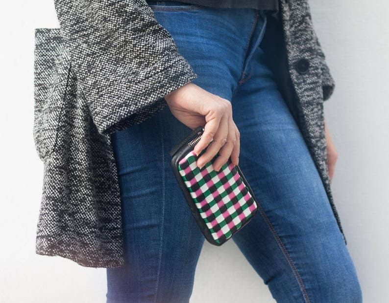 May include: A woman wearing a gray tweed coat and blue jeans is holding a black and white checkered wallet with a pink stripe.