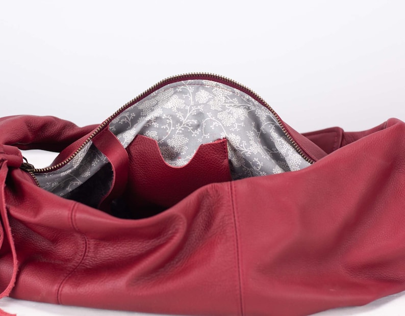 K&ouml;nnte beinhalten: Offene rote Lederhandtasche mit Rei&szlig;verschluss. Das Innenfutter ist ein grauer Stoff mit wei&szlig;em Blumenmuster. Eine kleine Tasche ist im Inneren der Tasche sichtbar.