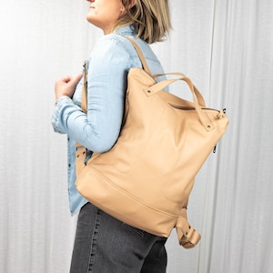 Puede incluir: Una mochila de cuero de color beige claro con un asa superior y correas ajustables. La bolsa tiene un cierre de cremallera y una forma rectangular. La mochila se lleva sobre los hombros.