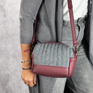 May include: A crossbody bag with a gray tweed center panel and burgundy leather accents. The bag has a rectangular shape with a quilted design on the tweed. The strap and bottom are burgundy, and the hardware is gold.