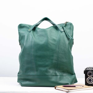 Peut inclure: Un grand sac fourre-tout en cuir vert avec une fermeture éclair et deux poignées supérieures. Le sac est posé sur une surface blanche avec un carnet et un stylo.