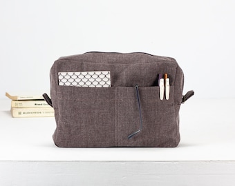 May include: A rectangular, brown fabric bag with multiple front pockets. A small notebook with a black and white pattern, two pens, and a pair of glasses are in the pockets. Two books are stacked on the left side of the bag.