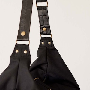 Op de afbeelding: Zwarte schoudertas met leren riemen en goudkleurige metalen hardware. De tas heeft een eenvoudig, minimalistisch ontwerp. De riemen zijn verstelbaar en de tas lijkt gemaakt van een stevig, waterbestendig materiaal.