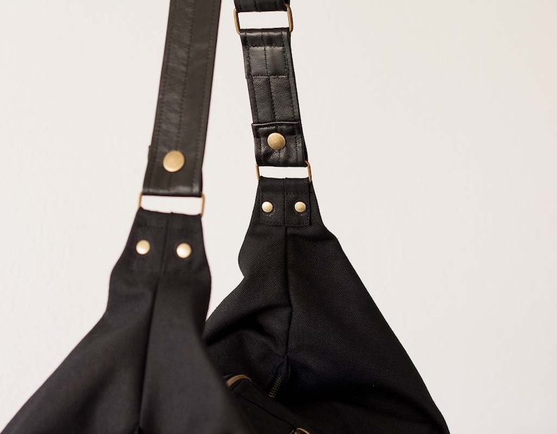 Op de afbeelding: Zwarte schoudertas met leren riemen en goudkleurige metalen hardware. De tas heeft een eenvoudig, minimalistisch ontwerp. De riemen zijn verstelbaar en de tas lijkt gemaakt van een stevig, waterbestendig materiaal.