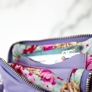 May include: A lavender zippered wallet with a floral lining. The wallet is open and has several cards inside.
