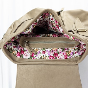 May include: A beige leather bag with a floral lining. The bag has a zipper closure and a flap with a buckle.