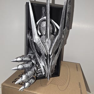 Puede incluir: Una escultura plateada y estilizada de la cabeza de una criatura con rasgos afilados y puntiagudos. La escultura est&aacute; montada sobre un marco rectangular negro y descansa sobre una caja de cart&oacute;n. La escultura tiene detalles intrincados y un acabado met&aacute;lico.