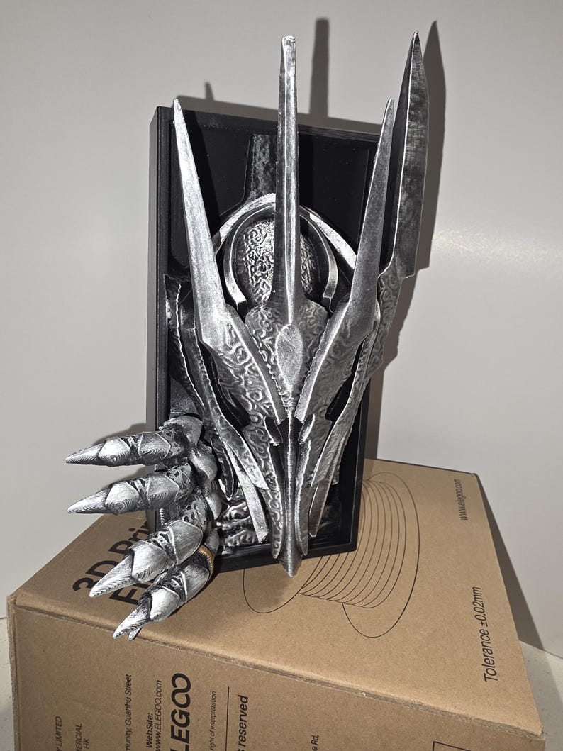 Puede incluir: Una escultura plateada y estilizada de la cabeza de una criatura con rasgos afilados y puntiagudos. La escultura est&aacute; montada sobre un marco rectangular negro y descansa sobre una caja de cart&oacute;n. La escultura tiene detalles intrincados y un acabado met&aacute;lico.