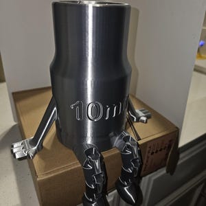Puede incluir: Una llave de vaso plateada, impresa en 3D, con brazos y piernas, sentada sobre una caja de cartón. La llave está grabada con el número "10mm". Los brazos y las piernas son articulados, lo que le da a la llave un aspecto antropomórfico juguetón.
