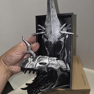 Puede incluir: Escultura detallada de una cabeza de drag&oacute;n plateada y negra con un cuerno largo y puntiagudo. La escultura est&aacute; montada sobre un marco rectangular negro. La boca del drag&oacute;n est&aacute; abierta, revelando afilados dientes.