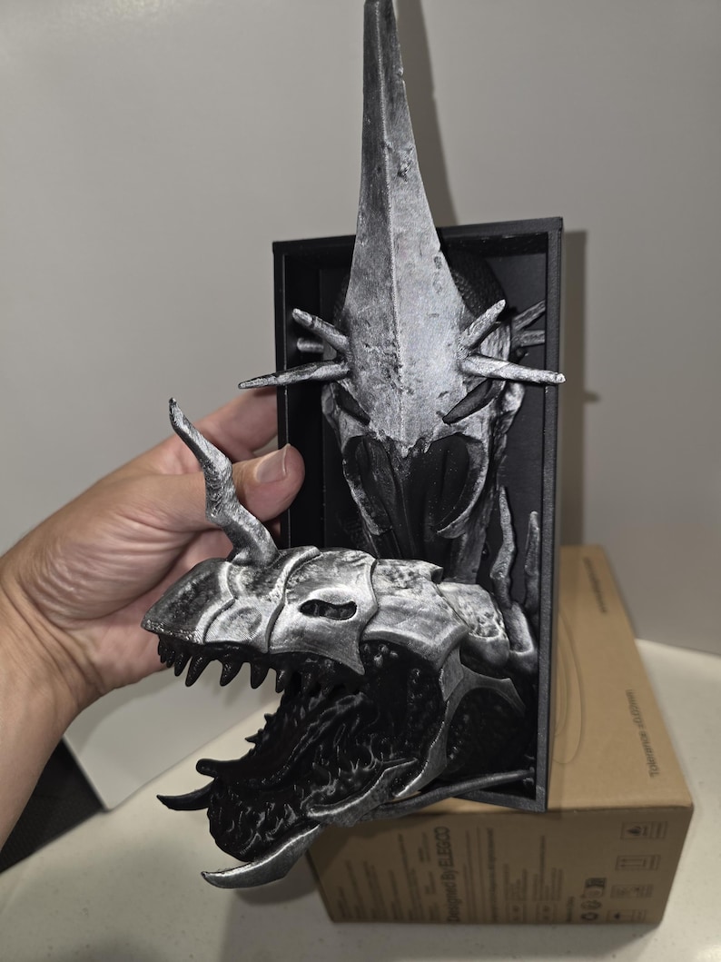 Puede incluir: Escultura detallada de una cabeza de drag&oacute;n plateada y negra con un cuerno largo y puntiagudo. La escultura est&aacute; montada sobre un marco rectangular negro. La boca del drag&oacute;n est&aacute; abierta, revelando afilados dientes.