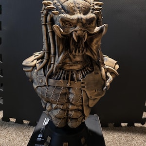 May include: A detailed bust of a Predator alien, featuring intricate details and a weathered bronze finish. The sculpture showcases the creature's iconic mandibles, dreadlocks, and armored chest. The bust is mounted on a black base.