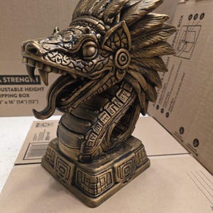 Op de afbeelding: Een goudkleurige sculptuur van een slangachtig wezen met een gevederde hoofdtooi en ingewikkelde details. De sculptuur heeft een vierkante basis met geometrische patronen en een lange, gespleten tong. Het is een decoratief item.