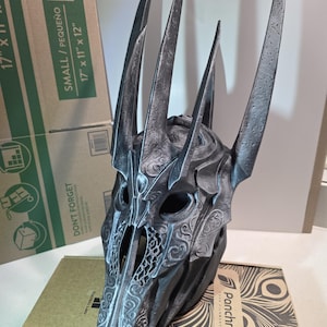 Könnte beinhalten: Ein detaillierter, dunkelgrauer Fantasy-Helm mit hohen, spitzen Stacheln. Der Helm weist komplizierte Muster und ein skelettartiges Design auf, das auf ein mittelalterliches oder gotisches Thema hindeutet. Das Produkt wird auf einer Box mit dem Text "DON'T FORGET" präsentiert.