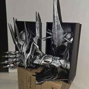 Puede incluir: Una detallada escultura plateada de la cabeza y la parte superior del cuerpo de una criatura, con rasgos afilados y puntiagudos. La escultura se exhibe en una caja negra, apoyada sobre una caja de cart&oacute;n. El dise&ntilde;o es intrincado y parece ser de tem&aacute;tica fant&aacute;stica.