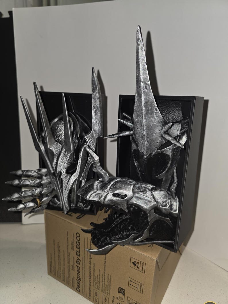 Puede incluir: Una detallada escultura plateada de la cabeza y la parte superior del cuerpo de una criatura, con rasgos afilados y puntiagudos. La escultura se exhibe en una caja negra, apoyada sobre una caja de cart&oacute;n. El dise&ntilde;o es intrincado y parece ser de tem&aacute;tica fant&aacute;stica.