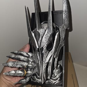 Puede incluir: Una escultura detallada de color plateado de la cabeza y la mano de un personaje de fantas&iacute;a, con dedos afilados en forma de garra y un anillo. La escultura tiene un fondo oscuro y est&aacute; sostenida en una mano. La escultura est&aacute; sobre una caja de cart&oacute;n.