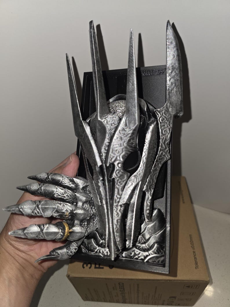 Puede incluir: Una escultura detallada de color plateado de la cabeza y la mano de un personaje de fantas&iacute;a, con dedos afilados en forma de garra y un anillo. La escultura tiene un fondo oscuro y est&aacute; sostenida en una mano. La escultura est&aacute; sobre una caja de cart&oacute;n.