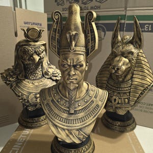 Puede incluir: Tres bustos egipcios dorados. El busto central representa a un faraón con una cofia de cobra. A los lados, bustos de Horus, el dios con cabeza de halcón, y Anubis, el dios con cabeza de chacal. Cada busto está sobre una base negra.