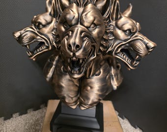 Puede incluir: Una escultura de color bronce de Cerbero, el perro de tres cabezas de la mitología griega. La escultura tiene una textura detallada y se asienta sobre una base negra. Las cabezas gruñen, con bocas abiertas y dientes visibles.