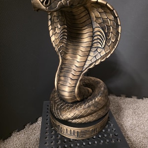 May include: A bronze-coloured cobra statue with a detailed, textured surface. The cobra is coiled and upright, with its hood flared. The statue sits on a black, geometric stand, and is likely a decorative item.