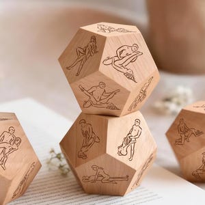 May include: Wooden dodecahedron dice with engraved illustrations of various sexual positions. The dice are made of light-colored wood and are arranged on a white surface. A mug is visible in the background.