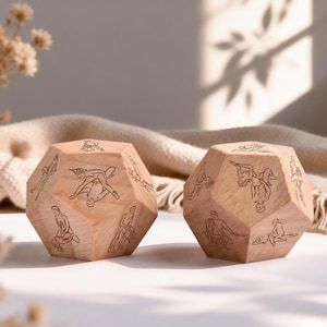 May include: Two wooden dodecahedron dice with etched illustrations of various positions. The dice are light brown and rest on a white surface with a beige blanket in the background. Dried flowers are in the foreground.