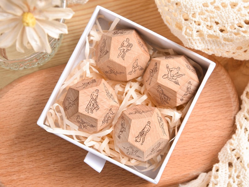 May include: A set of four wooden dice with various illustrated poses, presented in a white box with shredded paper. The dice are light brown and have black line drawings. The box sits on a wooden surface.