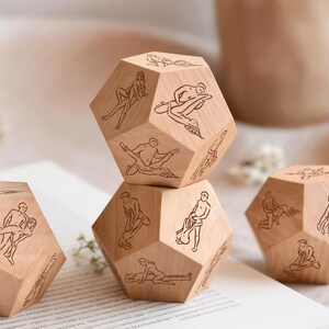 May include: Wooden dodecahedron dice with engraved illustrations of various sexual positions. The dice are made of light-colored wood and are arranged on a white surface. A mug is visible in the background.