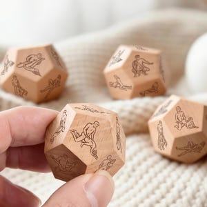 May include: Four wooden dodecahedron dice with etched illustrations of figures in various poses. The dice are light brown and rest on a cream-colored knitted surface. One die is held in a hand.