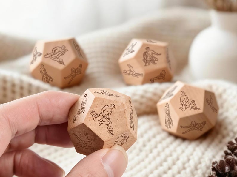 May include: Four wooden dodecahedron dice with etched illustrations of figures in various poses. The dice are light brown and rest on a cream-colored knitted surface. One die is held in a hand.