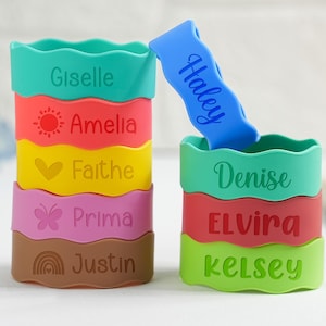 May include: Colorful silicone cup sleeves stacked with names and designs. The names Giselle, Amelia, Faithe, Prima, and Justin are on one stack. The other stack has Denise, Elvira, and Kelsey. A blue band with the name Haley is on top.