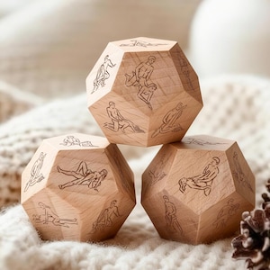 May include: Three wooden dodecahedron dice with etched illustrations of various sexual positions. The dice are made of light-colored wood and are arranged on a soft, white, textured surface. A pine cone is visible in the background.